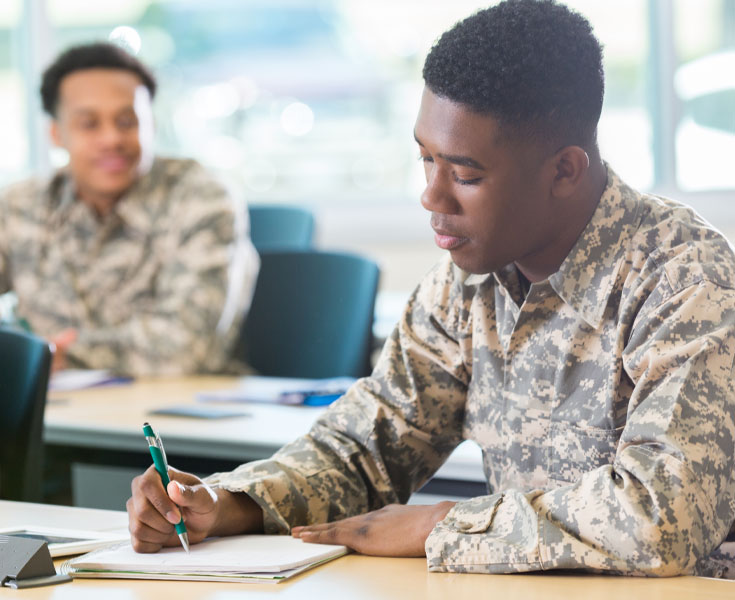 Military students writing on a pad of paper, while another student watches in background.
